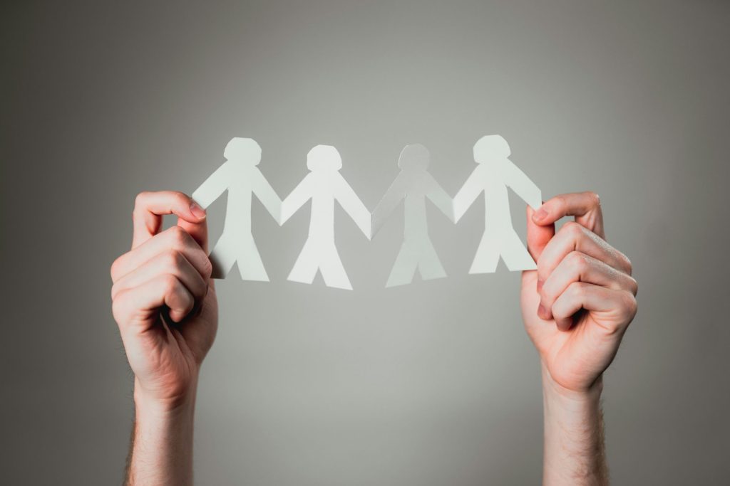 Two hands holding up a chain of white paper people cutouts against a neutral grey background, symbolizing community unity and collaborative support.